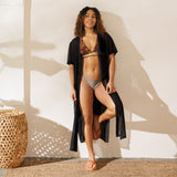 Woman in a bikini and cover-up standing in a sunlit room with a woven stool.
