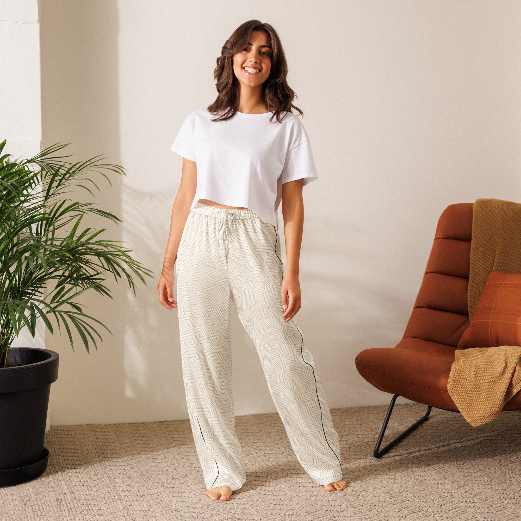 Woman wearing a white t-shirt and light-colored pants standing in a room with a plant and a chair.