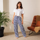 Woman wearing a white t-shirt and blue geometric patterned pants standing in a room with a plant and chair.
