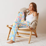 Woman sitting on a wooden chair wearing colorful floral pants and a beige cardigan.