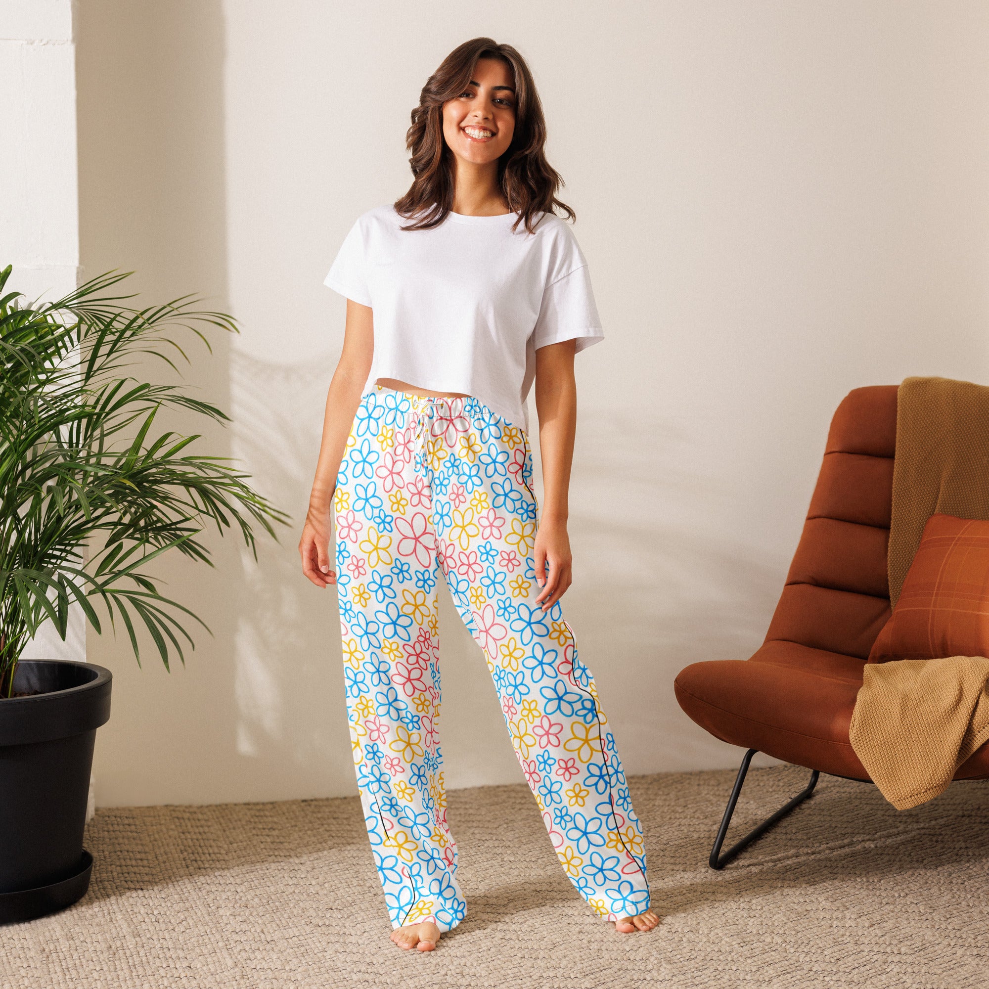 Woman wearing colorful floral pants and a white top in a room with a plant and chair.