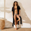 Woman in a bikini and cover-up standing in a sunlit room with a woven stool.
