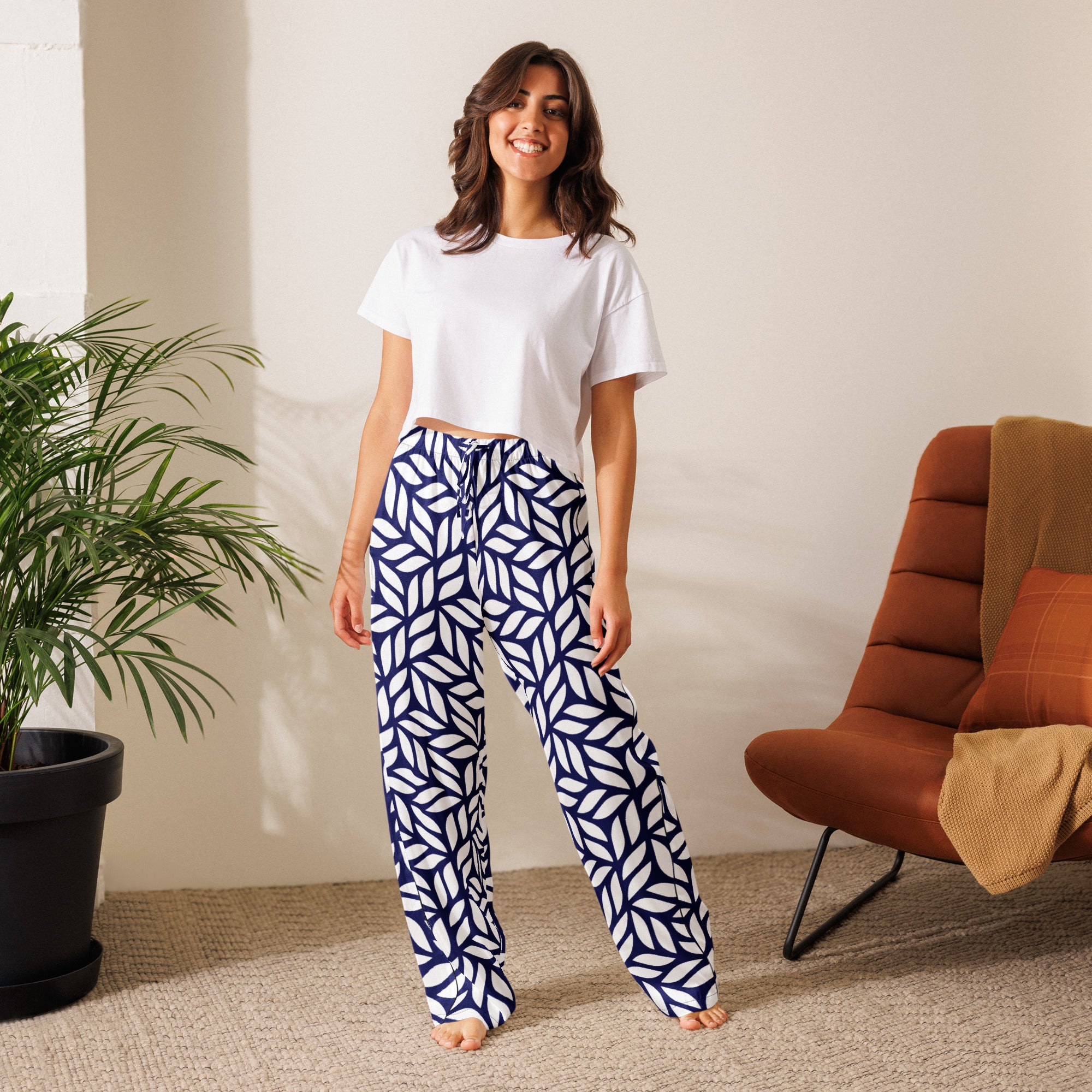 Woman wearing a white t-shirt and blue geometric patterned pants standing in a room with a plant and chair.