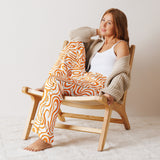 Woman sitting on a wooden chair wearing orange patterned pants and a beige cardigan.