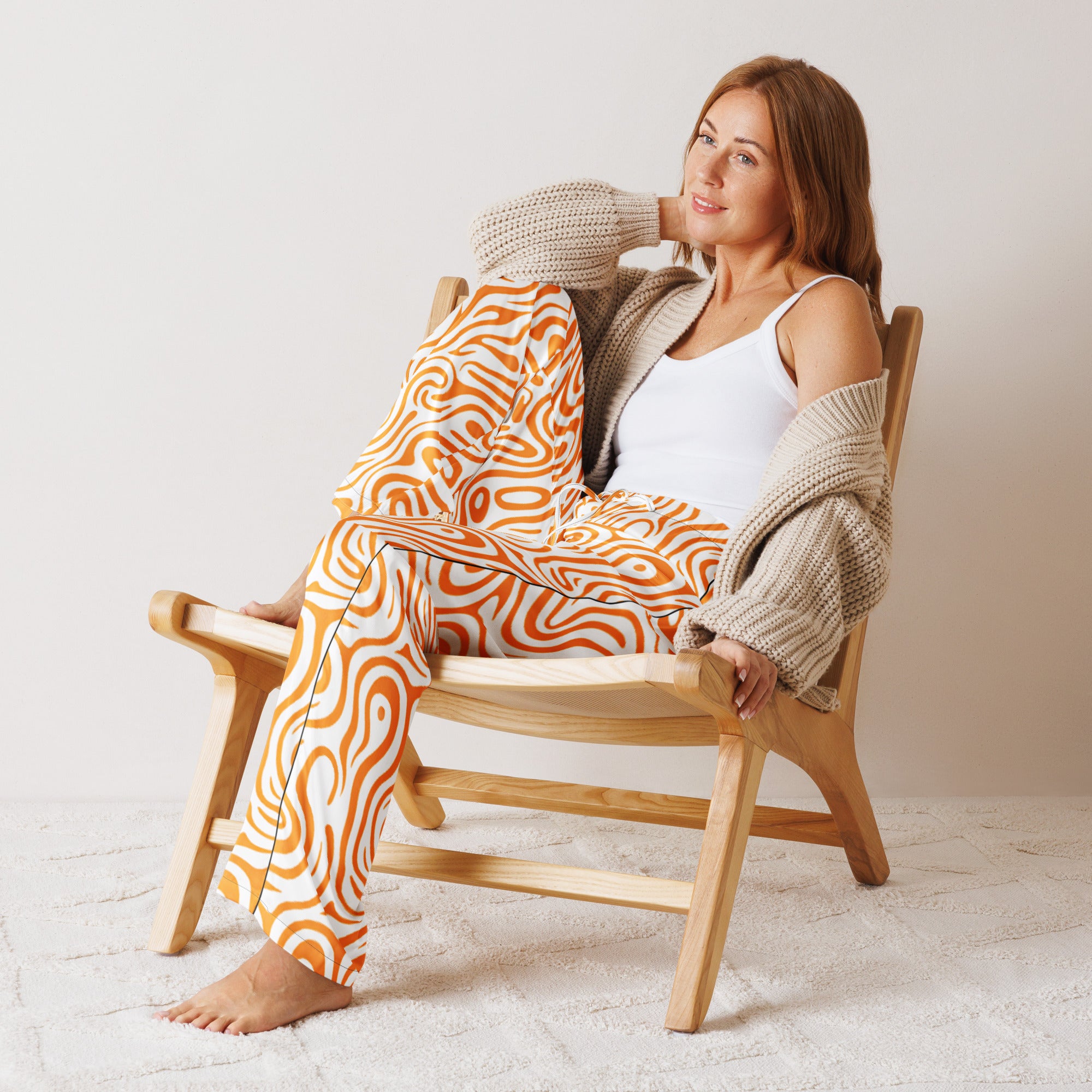 Woman sitting on a wooden chair wearing orange patterned pants and a beige cardigan.
