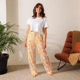 Woman wearing a white t-shirt and orange patterned pants standing in a room with a plant and chair.