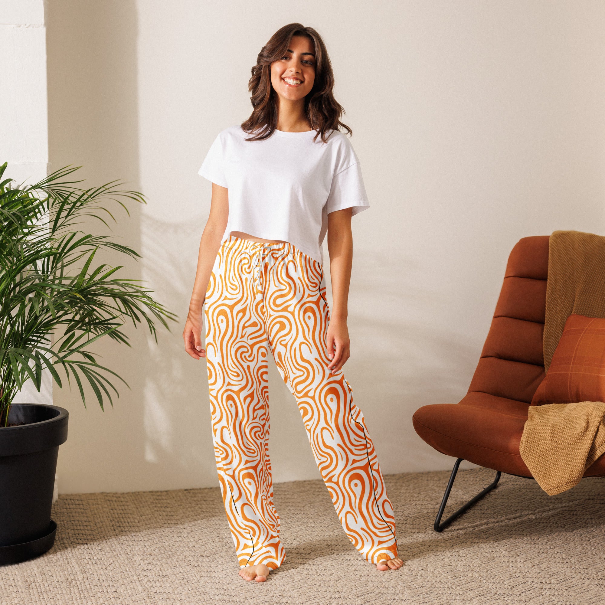 Woman wearing a white t-shirt and orange patterned pants standing in a room with a plant and chair.